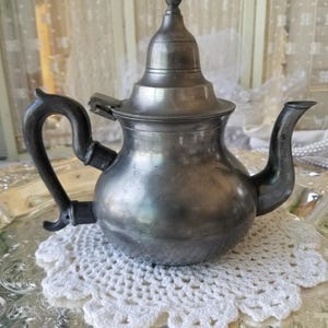 May include: A silver teapot with a domed lid and a decorative handle. The teapot is sitting on a white crocheted doily.