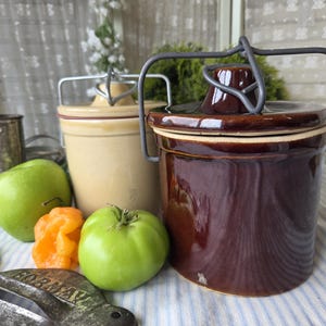 May include: Two cylindrical ceramic crocks with wire bail lids, one in a glossy dark brown glaze and the other in a matte beige finish. A green apple, an orange habanero pepper, a green tomato, and a metal tool are placed in front.