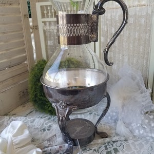 May include: A silver-plated glass coffee or tea pot with a decorative handle and a stand with three legs. The pot has a clear glass body and a silver-plated band around the middle.