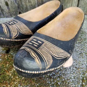 May include: Pair of black wooden clogs with intricate carved designs. The clogs have a light brown interior and a black exterior with a geometric pattern.
