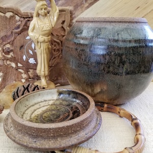May include: A collection of handcrafted pottery and decorative items. Includes a round, glazed ceramic pot with a bamboo handle, a small ceramic bowl, and a carved wooden figurine of a woman carrying a vessel on her head.