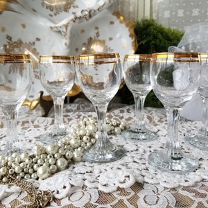 May include: Set of six clear glass wine glasses with a gold rim. The glasses are arranged on a white lace tablecloth with a string of pearls.