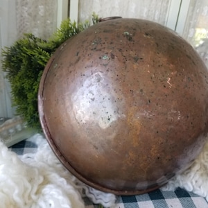 May include: A large, antique copper bowl with a rounded shape and a small handle. The bowl has a rich, aged patina with visible spots and imperfections, suggesting its vintage. The bowl is set against a backdrop of greenery and a checkered pattern.