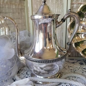 May include: A polished silver coffee pot with a curved spout and a decorative handle. The pot has a lid with a finial and a detailed band around the base. The pot is displayed with pearls and white lace.