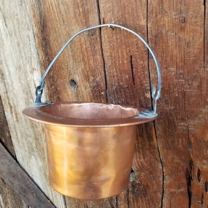 May include: A copper planter with a wire handle hanging on a wooden wall. The planter is empty and has a shiny, metallic finish.