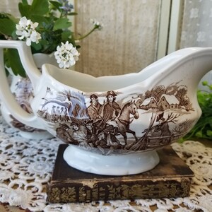 May include: A white porcelain gravy boat with a brown and white winter scene design. The scene features people on horseback and a snowy village.