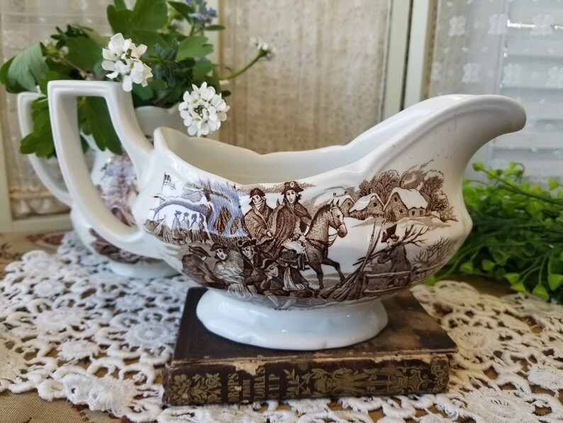 May include: A white porcelain gravy boat with a brown and white winter scene design. The scene features people on horseback and a snowy village.