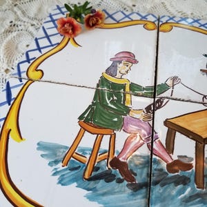 May include: Hand-painted ceramic tiles featuring a historical scene of a person working at a table. The tiles have a white background with a blue and yellow border. The person is wearing a green jacket, pink pants, and brown shoes.