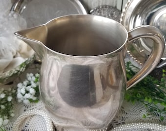 May include: A silver-colored pitcher with a curved handle and a spout. The pitcher is surrounded by other silver serving pieces, white lace, and a string of pearls. The pitcher has a shiny, reflective surface.