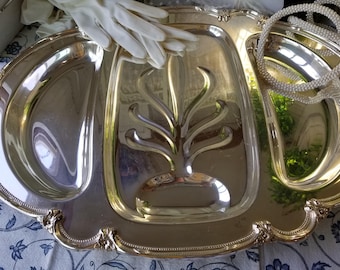 May include: A silver-colored serving tray with three compartments and ornate edges. The center section features a decorative, stylized tree design. A pair of white gloves and a beaded necklace are partially visible on the tray.