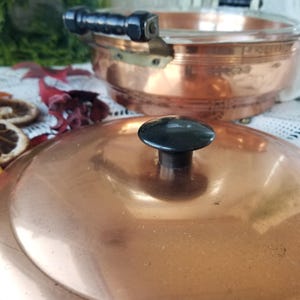 May include: A copper-colored cooking pot with a glass top and black handles. The lid, in the foreground, has a black knob. The pot is likely vintage, with a warm, inviting aesthetic. The pot is designed for cooking.