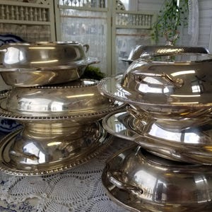 May include: A collection of stacked silver-plated serving dishes and lidded tureens with polished metallic surfaces, decorative beaded edges, and integrated side handles, arranged in a tiered formation on a white lace tablecloth.