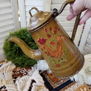 May include: An antique-style, olive-green metal teapot with a rooster and floral design. The teapot features a curved spout, a handle, and a lid with a small loop. The artwork includes red flowers and green leaves. The teapot is held by a hand.
