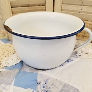 May include: A white enamelware chamber pot with a dark blue rim and a handle. The pot has a slightly distressed look, with some wear on the rim. It sits on a patchwork quilt with blue and white floral patterns.