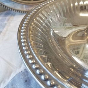 Puede incluir: Primer plano de una fuente de plata con borde de cuentas y dise&ntilde;o interior acanalado. La fuente es reflectante, mostrando el entorno. Otra fuente es parcialmente visible en el fondo.