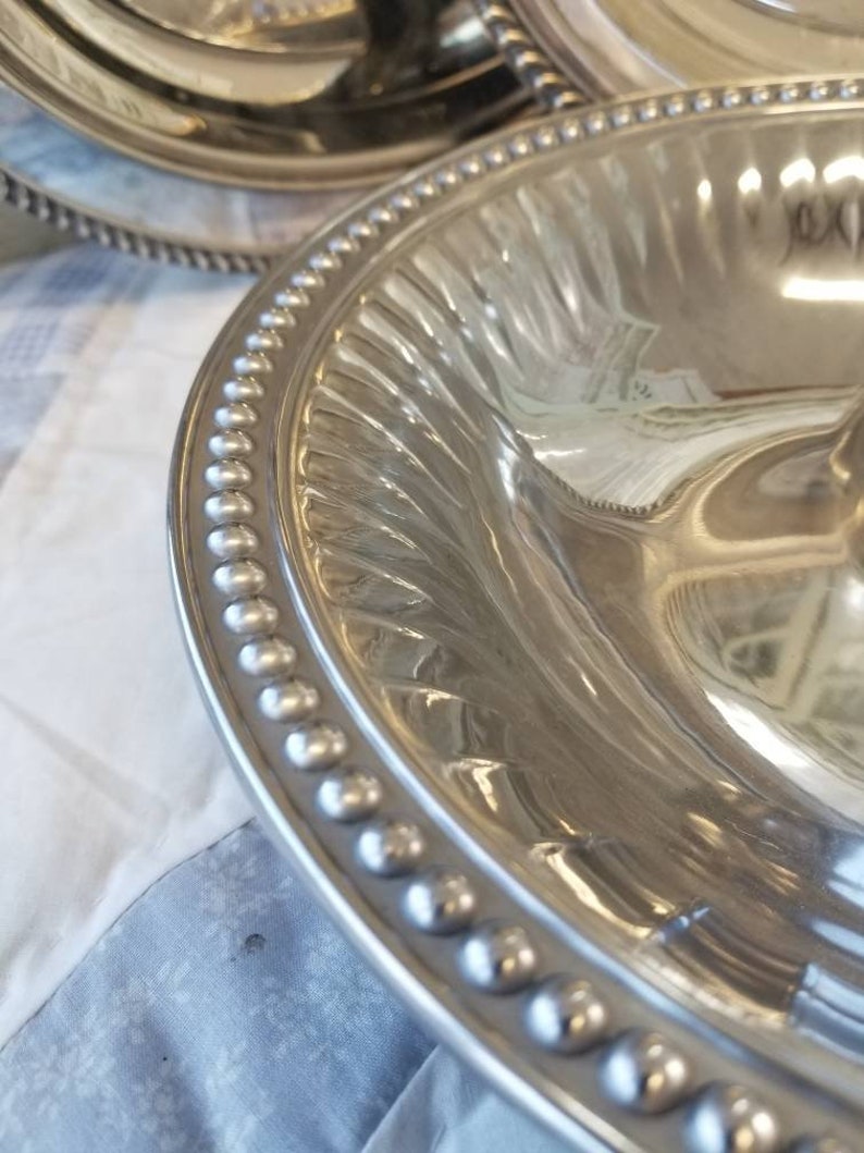 Puede incluir: Primer plano de una fuente de plata con borde de cuentas y dise&ntilde;o interior acanalado. La fuente es reflectante, mostrando el entorno. Otra fuente es parcialmente visible en el fondo.