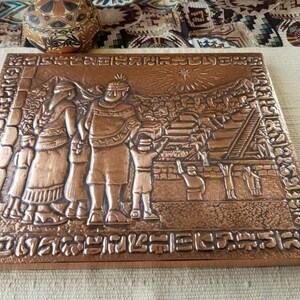 May include: A copper-colored, square relief sculpture depicting a historical scene with figures in traditional attire. The artwork features a detailed landscape with buildings and a sunburst design, framed by an ornate border.