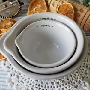 May include: A nested set of three white ceramic measuring bowls. The largest bowl has a handle and a pouring spout. The text on the bowls reads "1 cup 8floz" and "1/2 cup 4floz".