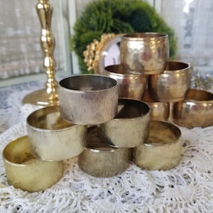 May include: A collection of gold-toned metal napkin rings. Some rings have a hammered texture, while others have a brushed finish. The rings are stacked on a white lace doily, suggesting a vintage or elegant dining theme.