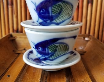 May include: Two white ceramic teacups with matching saucers. Each cup features a blue fish design with green accents. The cups are stacked on top of each other, resting on a bamboo tray. The background is a bamboo wall.
