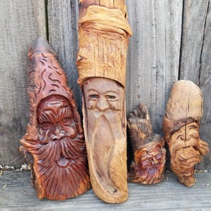 May include: Four hand-carved wooden sculptures of faces with long beards, each crafted from a distinct piece of natural wood. The carvings feature varied textures, ranging from smooth, light-toned wood to dark, deeply grooved, reddish-brown grain.