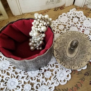 May include: A silver-colored, ornate jewelry box with a red velvet interior and a decorative lid. A cluster of faux pearls hangs over the edge. The box sits on a white lace doily.