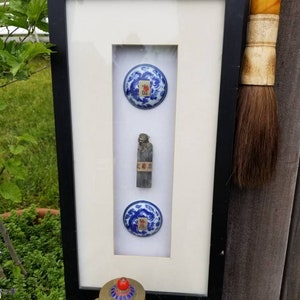 May include: Framed display featuring two blue and white porcelain circular objects with Chinese characters, and a small metal figurine. The frame is black with a white mat. A decorative brush with a wooden handle is visible.