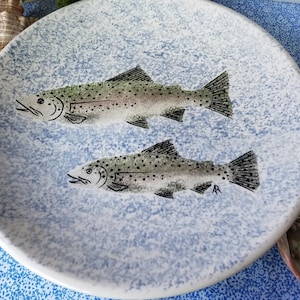 May include: A round, ceramic plate with a light blue speckled background. Two detailed illustrations of fish are centered on the plate. The fish are gray with black spots and fins. The plate has a white rim.