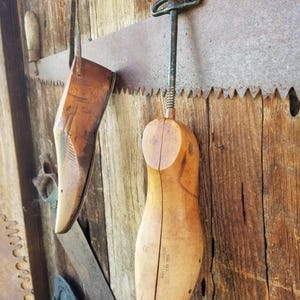 May include: A wooden shoe last and a shoe stretcher hang on a weathered wooden wall, alongside a rusty saw. The shoe last is a light brown color, and the stretcher has a green handle. The scene evokes a vintage workshop.