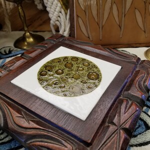 May include: A square, dark wood frame with carved details surrounds a white tile. The tile features a circular design with a mottled green and brown pattern. The frame is set against a backdrop of other decorative items.