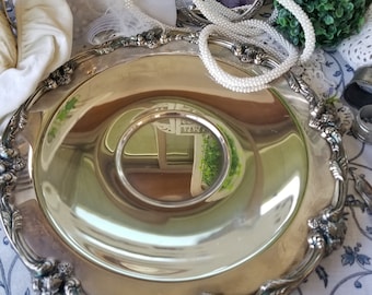 May include: A silver serving tray with an ornate, raised rim and a smooth, reflective surface. The tray is sitting on a white and blue floral tablecloth.