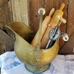 May include: A vintage brass coal scuttle filled with antique kitchen tools, including rolling pins and an egg beater. The scuttle sits on a blue and white quilt, with a rustic wooden background. The handle of the scuttle has a decorative face.