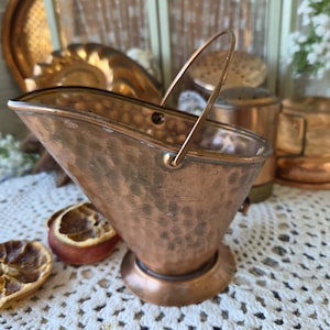 May include: A small, hammered copper coal scuttle with a wide, open top and a thin, arched metal handle. The vessel features a rounded base and a polished metallic finish, showcasing a textured, dimpled surface throughout the exterior body.