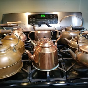 May include: A collection of seven copper tea kettles with various handle styles, including wood and metal, arranged on a black stovetop. The kettles feature rounded bodies, curved spouts, and lids, showcasing different shades of polished and aged copper.