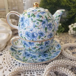 May include: A floral-patterned tea set featuring a teapot, teacup, and saucer. The set has a blue floral design with gold trim. The teapot has a gold knob on the lid. The teacup sits on the saucer, ready for tea time.
