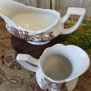 May include: A set of two white ceramic gravy boats with brown and white floral designs. The gravy boats are both empty and have a handle on the side.
