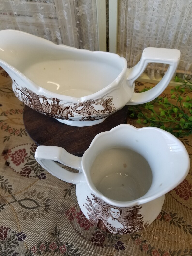 May include: A set of two white ceramic gravy boats with brown and white floral designs. The gravy boats are both empty and have a handle on the side.