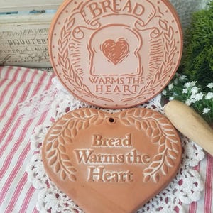 May include: Two terracotta bread-themed wall hangings. One is round with the word "BREAD" and "WARMS THE HEART" text, and a bread slice with a heart. The other is heart-shaped with the same text.