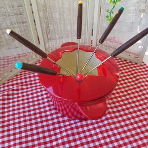 May include: Red fondue pot with six wooden handled dipping forks. The forks have colored tips: yellow, green, blue, and white. The pot is on a red and white checkered tablecloth.