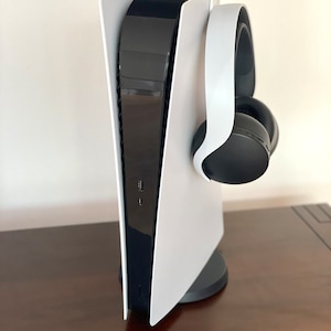 May include: A white PlayStation 5 console with a black headset hanging on the side. The console is standing on a dark brown wooden surface.