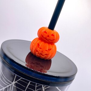May include: A black straw with two orange pumpkin-shaped toppers. The pumpkins have carved jack-o'-lantern faces. The straw is on top of a black and white spiderweb patterned tumbler.