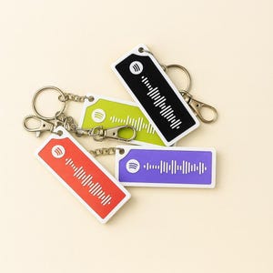 May include: Four rectangular keychains in red, green, black, and purple. Each keychain features a white Spotify logo and a white sound wave design. The keychains are attached to silver keyrings and clasps.