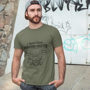 Puede incluir: Camiseta verde oliva con el texto "I Speak Fluent Dungeon Master" y bocadillos con frases como "Roll Initiative" y "Make a Dex save". La camiseta presenta un gráfico de dados y espadas.