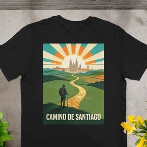 May include: Black t-shirt featuring a colorful graphic of a person hiking the Camino de Santiago. The design includes a path leading to a cathedral under a rising sun, with the text "CAMINO DE SANTIAGO" below.