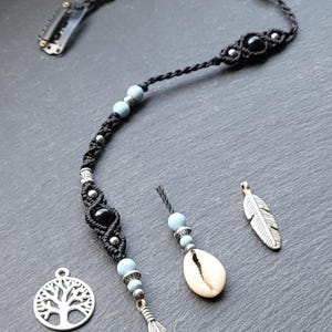 Puede incluir: Un cordón negro con cuentas azules y un dije de pluma de plata. El cordón tiene un clip de plata en un extremo y un dije de árbol de plata en el otro extremo. También hay un pequeño dije de concha blanca.