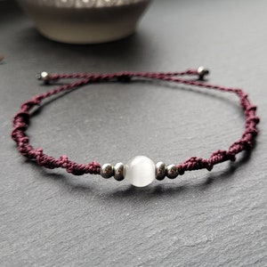 Könnte beinhalten: Ein handgefertigtes Armband mit einem zentralen weißen Edelstein und silberfarbenen Perlen, auf einer dunkelburgunderfarbenen Kordel aufgereiht. Die Kordel ist geknotet und das Armband ist verstellbar. Es ist ein Schmuckstück.