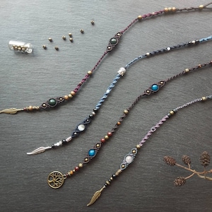 Peut inclure: Cinq colliers en cordon macramé faits à la main avec différentes perles de pierres précieuses, des breloques en métal et des plumes. Les colliers sont présentés sur une surface gris foncé.