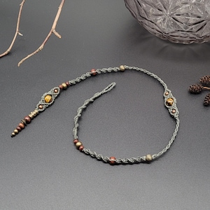 Könnte beinhalten: Eine graue Makramee-Halskette mit braunen und goldenen Perlen und einem Anhänger aus Tigerauge.