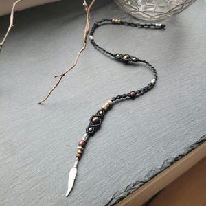 Peut inclure: Un collier en cordon noir avec des perles brunes et dorées et un pendentif en plume argentée. Le collier est noué avec un motif macramé.