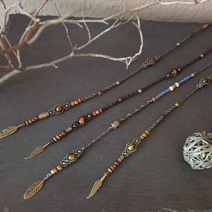 Op de afbeelding: Vier macramé haarwraps met bronzen veerkettingen. Elke wrap heeft een uniek ontwerp met bruine, gouden en blauwe kralen en een centrale tijgeroogsteen. Gepresenteerd op een donkere ondergrond met een decoratieve bal.